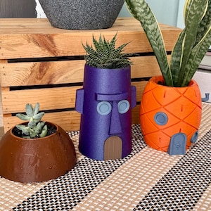 May include: Three decorative planters, each holding a succulent or plant. The planters are shaped like a brown dome, a purple Easter Island head, and an orange pineapple. The planters sit on a striped beige and black surface.