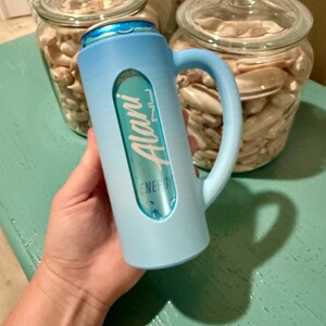 Puede incluir: Un enfriador de latas azul claro con asa, que sostiene una lata de bebida energ&eacute;tica Alani Nu. El enfriador tiene un recorte para mostrar la etiqueta de la bebida. Dos tarros de cristal llenos de conchas marinas est&aacute;n en el fondo.
