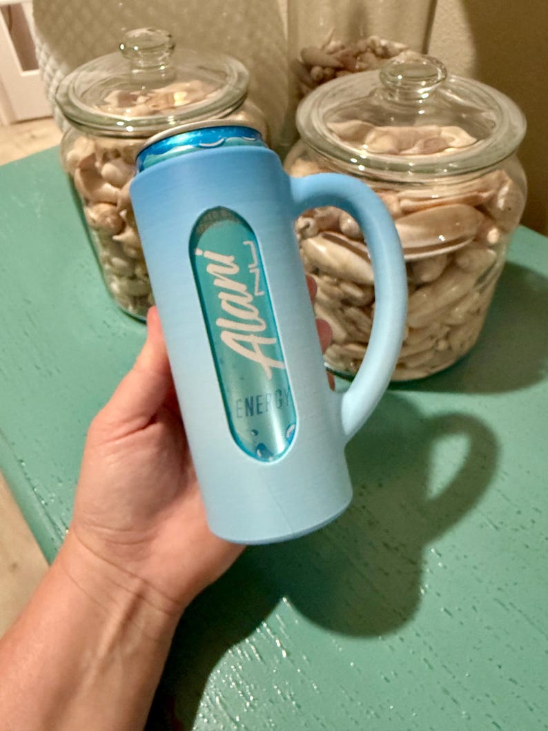 Puede incluir: Un enfriador de latas azul claro con asa, que sostiene una lata de bebida energ&eacute;tica Alani Nu. El enfriador tiene un recorte para mostrar la etiqueta de la bebida. Dos tarros de cristal llenos de conchas marinas est&aacute;n en el fondo.