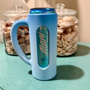 Puede incluir: Un enfriador de latas azul claro con asa, que contiene una lata turquesa de bebida energ&eacute;tica Alani Nu. El enfriador tiene un recorte para mostrar la etiqueta de la lata. El fondo incluye frascos de vidrio llenos de conchas marinas.