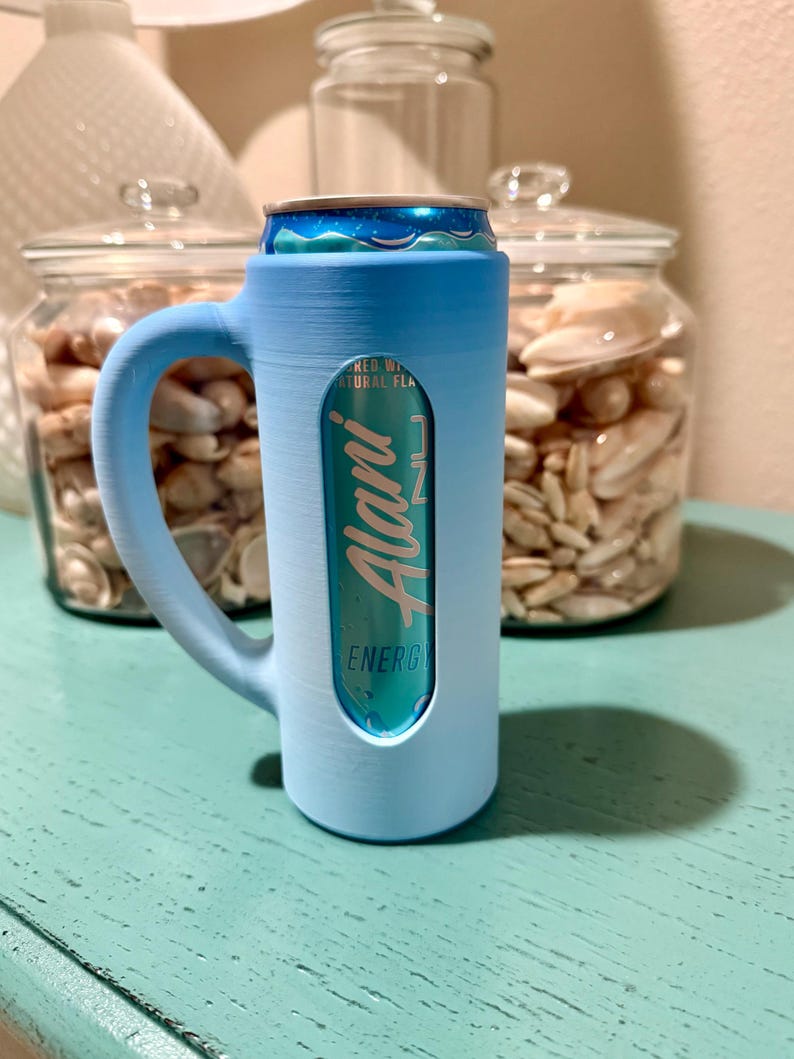 Puede incluir: Un enfriador de latas azul claro con asa, que contiene una lata turquesa de bebida energ&eacute;tica Alani Nu. El enfriador tiene un recorte para mostrar la etiqueta de la lata. El fondo incluye frascos de vidrio llenos de conchas marinas.