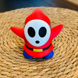 May include: A red and white 3D printed figurine of a character from the Super Mario franchise. The character is wearing a red hooded cloak, a white mask with black eyes, a yellow belt, and blue pants.