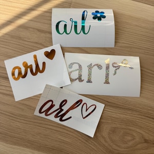 May include: Several white vinyl stickers with the word "arl" in various colors and fonts. One sticker has a teal font with a teal flower, another has a gold font with a heart, a third has a glittery font with a bow, and the last has a red font with a heart.