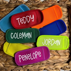 May include: A collection of colorful, personalized drink sleeves in various shades including red, orange, blue, green, and purple. Each sleeve features a name in white script: Teddy, Coleman, Jordan, and Penelope. The sleeves are displayed in a woven basket.