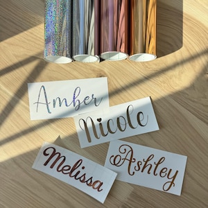 May include: Rolls of metallic vinyl in silver, gray, rose gold, and gold are displayed above four name decals. The decals read "Amber", "Nicole", "Melissa", and "Ashley" in various metallic colors.
