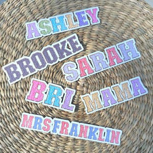 May include: A collection of colorful embroidered name patches, including "ASHLEY," "BROOKE," "SARAH," "BRL," "MAMA," and "MRS FRANKLIN." Each patch features a white border and a variety of pastel colors, set against a woven background.