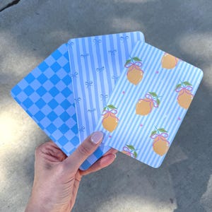 May include: Three blue and white patterned cards. One card has a checkered pattern, another has vertical stripes and bows, and the third has vertical stripes with lemons and bows. The cards are held in a hand.