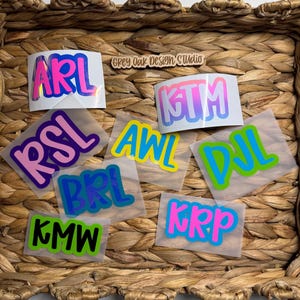 May include: A collection of colorful vinyl stickers with various three-letter acronyms. The stickers feature a gradient or solid color fill with a contrasting outline. The text "Grey Oak Design Studio" is visible in the background.