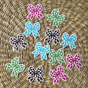 May include: A collection of colorful bow-shaped stickers with a checkered pattern. The stickers feature various colors, including pink, blue, green, and black, with white outlines. They are arranged on a woven, textured background.