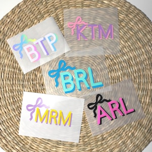 May include: Five personalized iron-on transfers with initials and bow designs. The initials are in various colors, including blue, pink, purple, and yellow. The bows are in coordinating colors. The transfers are on a clear backing.
