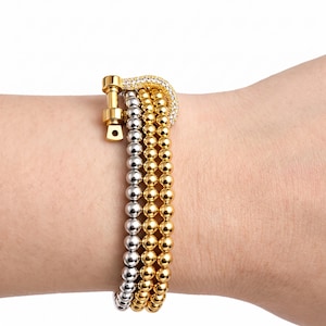 May include: A multi-strand beaded bracelet featuring two rows of gold-toned beads and one row of silver-toned beads. The bracelet is secured with a gold-toned horseshoe-shaped clasp that is embellished with small, clear, sparkling stones.