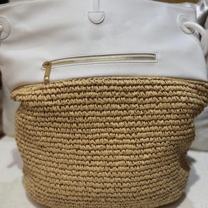 K&ouml;nnte beinhalten: Eine wei&szlig;e und beige Tragetasche. Der obere Teil ist wei&szlig; mit einem goldenen Rei&szlig;verschluss. Die untere H&auml;lfte besteht aus gewebtem beigem Material. Die Tasche hat wei&szlig;e Griffe und einen wei&szlig;en Riemenverschluss.