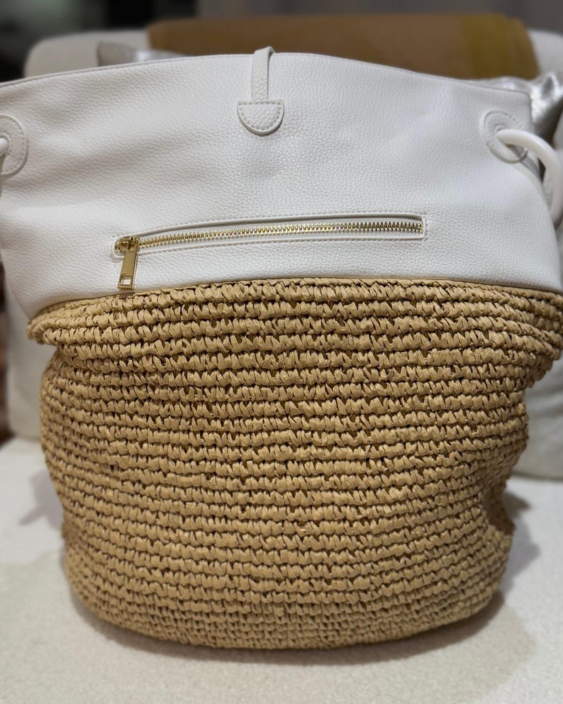 K&ouml;nnte beinhalten: Eine wei&szlig;e und beige Tragetasche. Der obere Teil ist wei&szlig; mit einem goldenen Rei&szlig;verschluss. Die untere H&auml;lfte besteht aus gewebtem beigem Material. Die Tasche hat wei&szlig;e Griffe und einen wei&szlig;en Riemenverschluss.