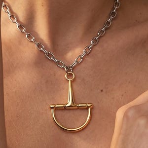 May include: A necklace featuring a thick silver-toned chain with a large, polished gold-toned horse bit pendant. The pendant is suspended from a circular ring attached to the center of the chain, showcasing a smooth, metallic, equestrian-inspired design.