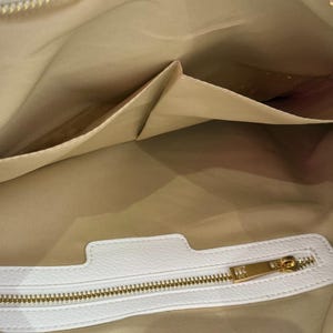 K&ouml;nnte beinhalten: Innenansicht einer wei&szlig;en Lederhandtasche mit goldenem Rei&szlig;verschluss und mehreren Taschen. Das Innere der Tasche ist hellbraun und der Rei&szlig;verschluss ist goldfarben. Die Tasche scheint neu und unbenutzt zu sein.