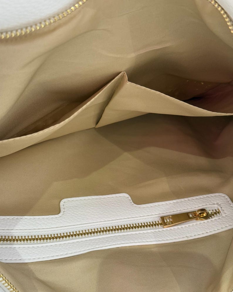 K&ouml;nnte beinhalten: Innenansicht einer wei&szlig;en Lederhandtasche mit goldenem Rei&szlig;verschluss und mehreren Taschen. Das Innere der Tasche ist hellbraun und der Rei&szlig;verschluss ist goldfarben. Die Tasche scheint neu und unbenutzt zu sein.