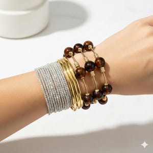 Puede incluir: Una colección de pulseras de varios estilos. Hay múltiples pulseras finas de diamantes de imitación plateados, varias pulseras doradas y un conjunto de pulseras de cuentas marrones con detalles dorados. Las pulseras están apiladas en una muñeca.