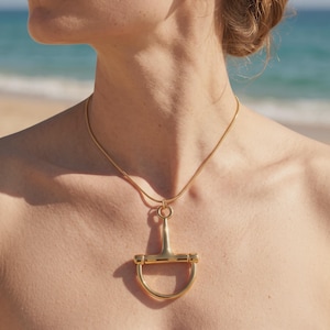 May include: A gold-toned necklace featuring a smooth, polished metal pendant shaped like a horse bit. The pendant is suspended from a thin, flexible gold-toned chain that rests around the neck. The metal has a reflective, high-shine finish.