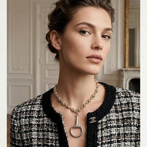 May include: A woman wearing a gold-toned chain necklace with a silver bit pendant. She is wearing a black and white tweed jacket with black trim and gold buttons. The background is a room with white walls and a gold-framed mirror.