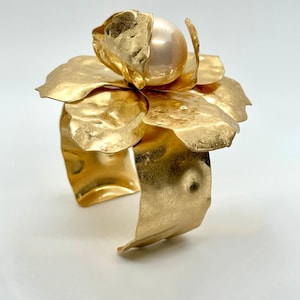 May include: Gold-tone cuff bracelet featuring a large, textured flower design. The flower has multiple petals and a large, round, off-white pearl at its center. The bracelet has a hammered metal finish and a wide band.