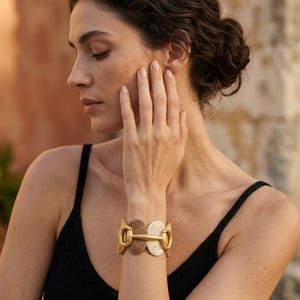 May include: A gold-toned bracelet with a woven design. The bracelet features two circular woven sections in beige and cream, connected by a gold-colored clasp. The model is wearing a black tank top.