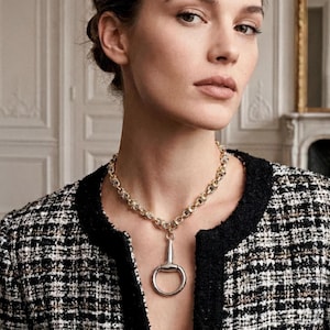 May include: A gold-toned chain necklace featuring a prominent silver-toned horse bit pendant. The necklace is worn around the neck, resting against a black and white tweed jacket with a textured pattern and black trim along the front edges.