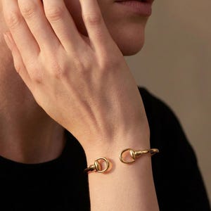 May include: A gold-toned metal cuff bracelet featuring a polished finish and a horsebit-inspired design. The bracelet is open-ended, with two rounded, loop-shaped terminals that face each other, creating a sleek and continuous metallic aesthetic.