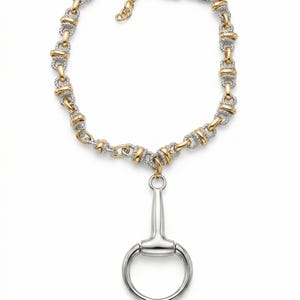 May include: A necklace featuring a gold and silver chain with a horse bit pendant. The chain is composed of alternating gold and silver links. The pendant is a silver horse bit. The necklace is displayed on a white background.