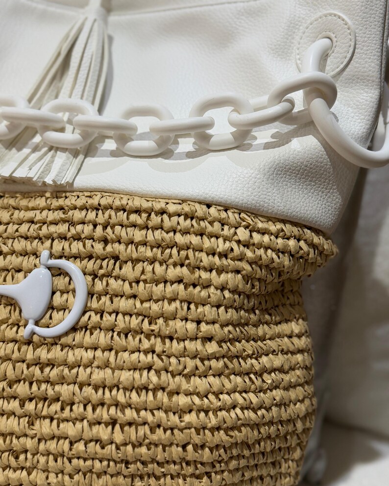 K&ouml;nnte beinhalten: Eine wei&szlig;e Handtasche mit gewebtem Strohboden. Die Tasche hat einen wei&szlig;en Kettenhenkel und eine wei&szlig;e Quaste. Die Tasche hat einen wei&szlig;en Verschluss und einen wei&szlig;en Griff. Die Tasche ist im Shopper-Stil.