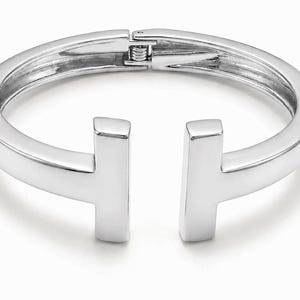 May include: A polished silver-tone hinged bangle bracelet featuring a T-shaped open design. The bracelet has a smooth, reflective metallic finish and a secure clasp mechanism at the top, creating a clean, continuous circular silhouette.