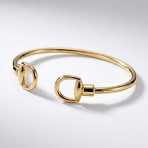 May include: A gold-colored open cuff bracelet. The bracelet features a horse bit design on each end. The metal has a polished finish, reflecting light. The bracelet is displayed on a white surface.