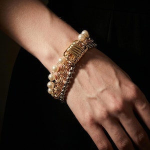 May include: A multi-strand bracelet featuring a row of white pearls, a gold-toned chain, and a silver-toned chain, all connected by a central gold-toned metal clasp with a series of horizontal wire wraps across the front surface.