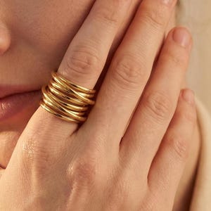 May include: A gold-colored ring with a multi-layered design. The ring is made of several intertwined bands, creating a textured and eye-catching appearance. The ring is worn on a finger, with the hand in the foreground.