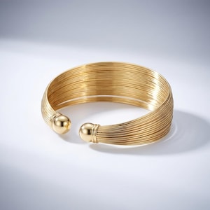 May include: A gold-colored cuff bracelet. The bracelet is made of numerous thin, parallel wires, giving it a textured appearance. The ends of the bracelet have rounded, solid gold accents. The open design allows for easy wear. This jewelry piece is suitable for various occasions.