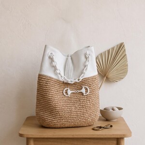 K&ouml;nnte beinhalten: Eine wei&szlig;e und beige Handtasche mit Kettenriemen und Quaste. Der obere Teil ist wei&szlig;, w&auml;hrend der untere Teil aus gewebtem beigem Material besteht. Ein dekorativer Bit-Verschluss und Kettendetails erg&auml;nzen das Design der Tasche. Die Tasche steht auf einem Holztisch.