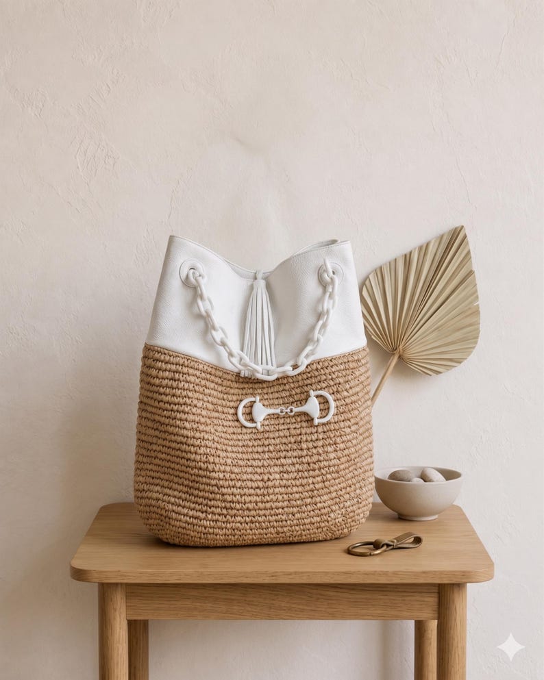 K&ouml;nnte beinhalten: Eine wei&szlig;e und beige Handtasche mit Kettenriemen und Quaste. Der obere Teil ist wei&szlig;, w&auml;hrend der untere Teil aus gewebtem beigem Material besteht. Ein dekorativer Bit-Verschluss und Kettendetails erg&auml;nzen das Design der Tasche. Die Tasche steht auf einem Holztisch.
