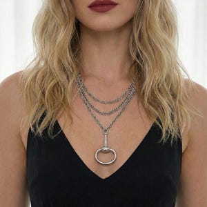 May include: Silver-toned necklace featuring three layered chains. The necklace has a large, circular pendant with a smaller, elongated piece attached. The jewelry is worn against a black top.