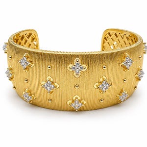 May include: A wide gold cuff bracelet featuring a textured, brushed finish. The surface is decorated with multiple four-petaled floral motifs, each set with small clear stones, interspersed with small gold dots. The interior has a lattice pattern.