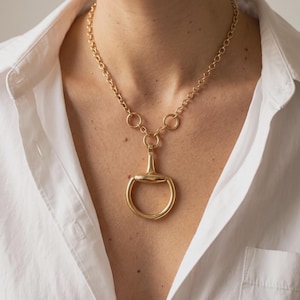 May include: A gold-toned necklace featuring a polished horsebit pendant suspended from a thick, round-link chain. The pendant consists of a horizontal bar connected to a circular ring, creating a classic equestrian-inspired design in a metallic finish.