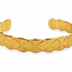May include: A gold-toned cuff bracelet featuring a repeating diamond-shaped quilted pattern across the exterior surface. The metal band has a wavy, scalloped edge along the top and bottom, with an open back design for sliding onto the wrist.
