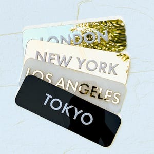 May include: Four rectangular plaques with city names: London, New York, Los Angeles, and Tokyo. The London plaque is mirrored, New York is gold, Los Angeles is white, and Tokyo is black. Each plaque has the city name in a contrasting color.