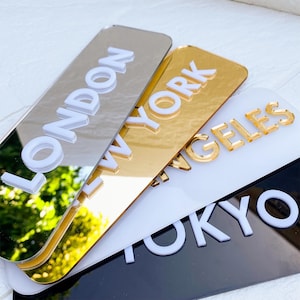 Op de afbeelding: Een verzameling rechthoekige, reflecterende plaquettes met verhoogde letters van stadsnamen. De plaquettes zijn in zilver, goud, wit en zwart. De steden zijn Londen, New York, Los Angeles en Tokio.