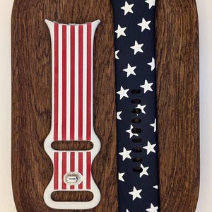 May include: Two patriotic Apple Watch bands. One features red and white stripes, the other has white stars on a navy blue background. Both bands are displayed on a wooden oval.