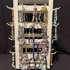 May include: A white wooden jewelry display stand with multiple hooks holding various beaded bracelets. The bracelets feature diverse colors, including shades of blue, purple, green, orange, and clear beads, with some incorporating small metal charms.