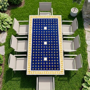 May include: A rectangular outdoor dining table with a blue and yellow geometric tile top featuring three white star-shaped accents, surrounded by eight light grey chairs with armrests, arranged in a symmetrical layout on a green lawn.