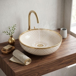 May include: A round ceramic vessel sink with a white and gold lattice pattern and a gold rim. A tall, curved gold tap dispenses water into the basin. The sink sits on a dark wood countertop alongside a small gold vase, a bar of soap, and a folded cloth.