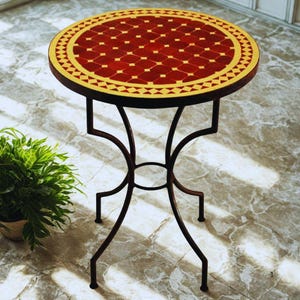 Pode incluir: Uma mesa lateral redonda em mosaico com um tampo de azulejos vermelhos e amarelos e uma base de metal castanho escuro. A mesa tem um padrão decorativo e é adequada para uso interior ou exterior. A mesa tem cerca de 61 cm de diâmetro.