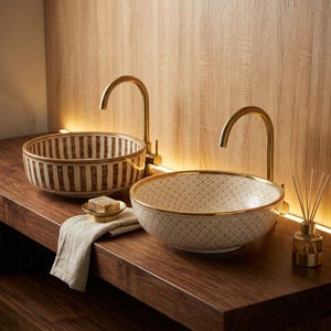 May include: Two round ceramic vessel sinks with gold-toned taps. The left sink features a brown and white vertical striped pattern, while the right sink has a gold-outlined lattice design. A small soap dish with a bar of soap and a reed diffuser sit nearby.