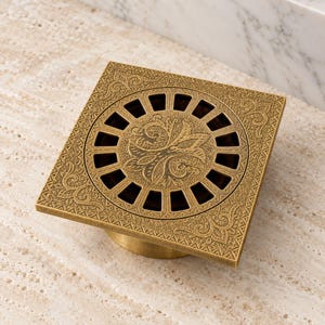 May include: A square brass floor drain cover featuring an intricate embossed pattern on the border and a central circular grate with a decorative floral motif. The metal surface has a brushed antique finish with visible dark recessed details.