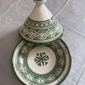 Puede incluir: Una olla de tagine de cerámica blanca y verde con tapa cónica y base redonda. La olla presenta intrincados patrones geométricos en verde, con un borde verde a juego. El tagine está abierto, revelando el interior.